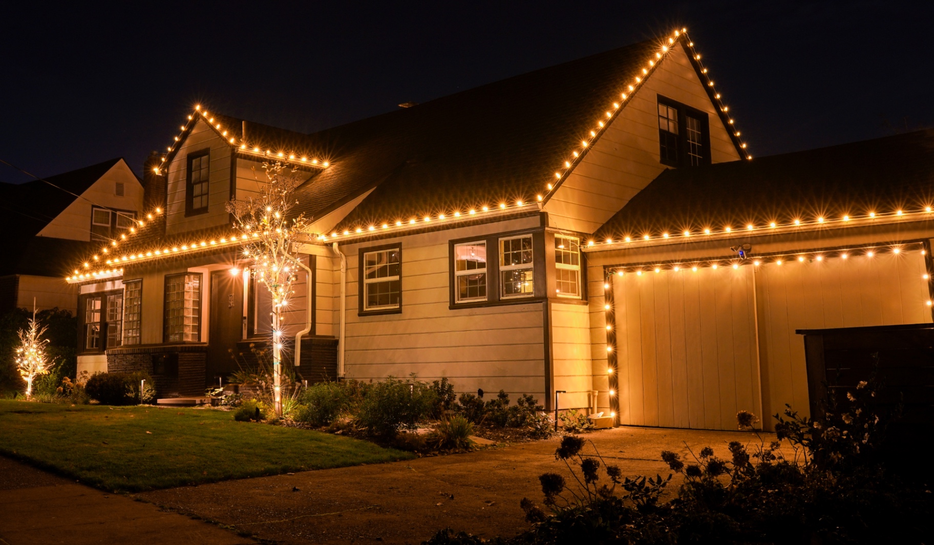 Craftsman home fully outlined in warm-white C9 Christmas lights across roofline, garage and wrapped front trees